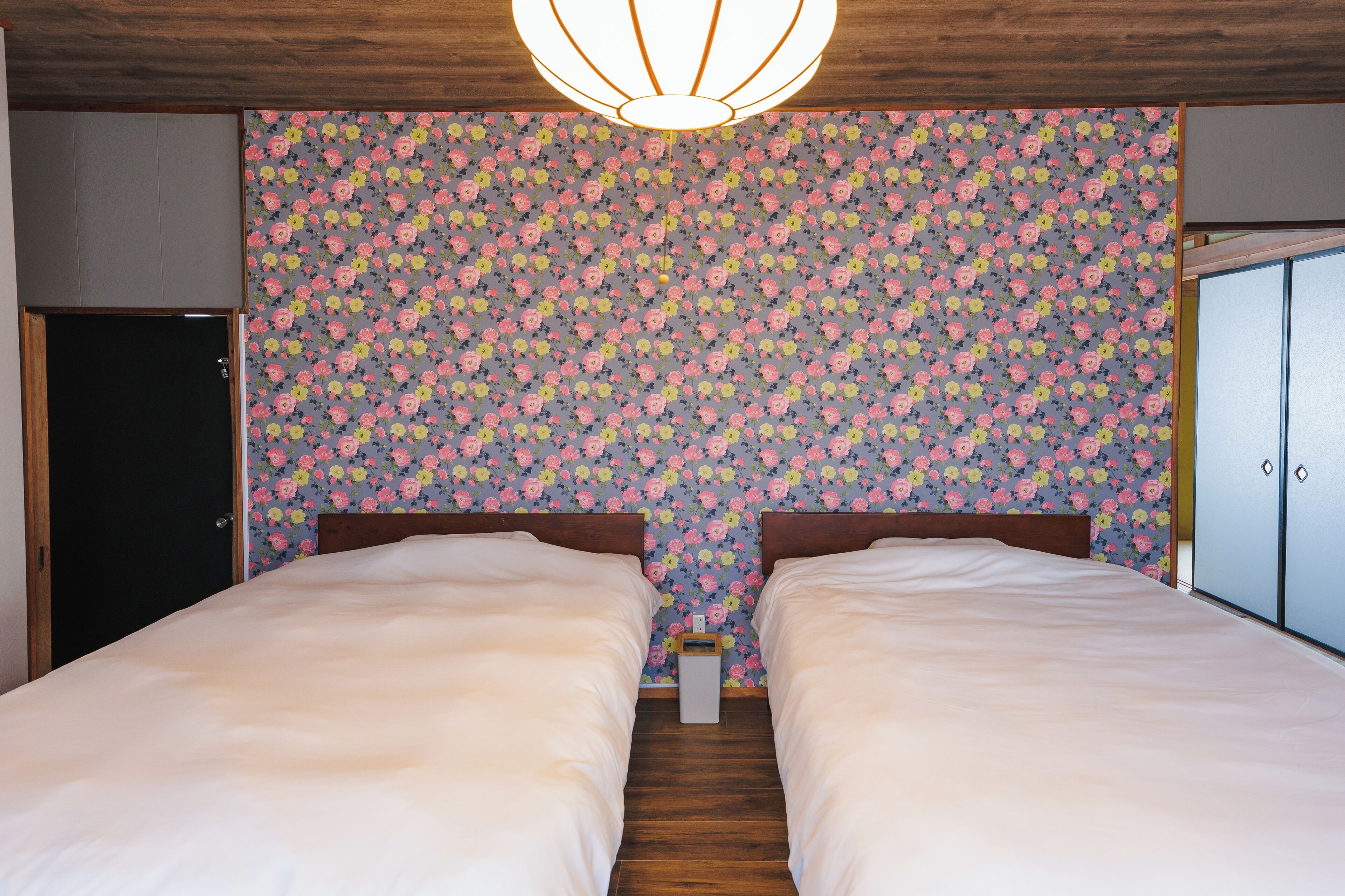 A bedroom with two beds, wooden flooring, a floral wallpaper, and a ceiling light fixture.