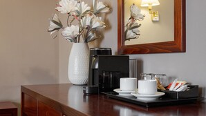Een houten toonbank met een koffiezetapparaat, bekers en een schaal met snacks. Op de toonbank staat een vaas met bloemen, en er is een wandlamp te zien.