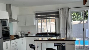 A modern kitchen with white cabinets, a stainless steel range hood, and a built-in oven. There is a dining area with a table and two chairs, and a view of a swimming pool through a sliding glass door. - Charming 3 bed villa 10 mins to the nearest & most beautiful beach in Mauritius (Morcellement Swan, Pereybere, Grand Baie)