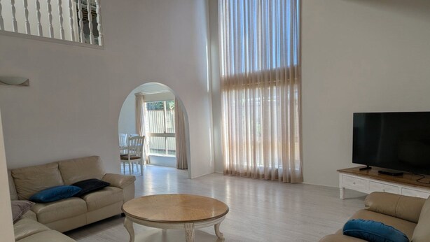Amplia sala de estar con televisor grande, una mesa redonda y dos sofás. Una ventana grande con cortinas y un arco de entrada que conduce a otra habitación.