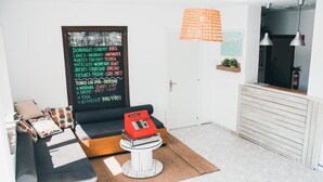 Un espace de vie compact comprenant un canapé, une petite table sur laquelle se trouve un objet rouge, un tableau noir servant de menu, et un coin cuisine avec un luminaire suspendu.