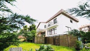 Une maison à deux étages dotée d’un balcon, entourée d’une clôture en bois et d’une végétation luxuriante.