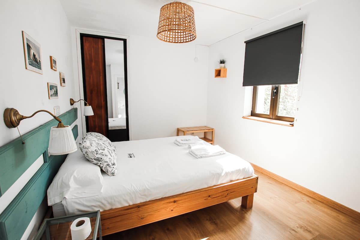 Een netjes opgemaakt bed met witte lakens, een houten hoofdbord, een bedlampje en een raam met zonwering.