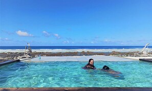 Área de piscina com água azul cristalina, uma pessoa e um cachorro, e vista para o oceano.