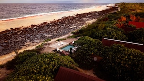 Vista aérea de un resort frente a la playa, con zona de piscina, sillas de descanso y un litoral rocoso.