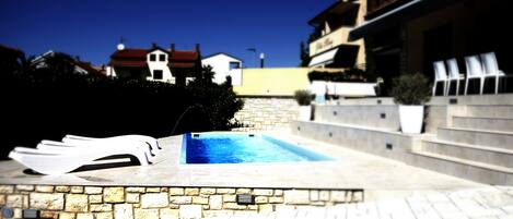 A swimming pool with stone tiles, a stone wall, and a white building in the background.