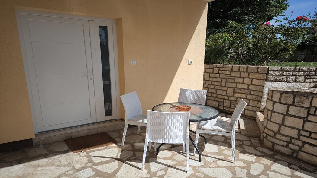 Un patio exterior con una mesa de vidrio y sillas de mimbre blanco, una puerta blanca y una pared de piedra.