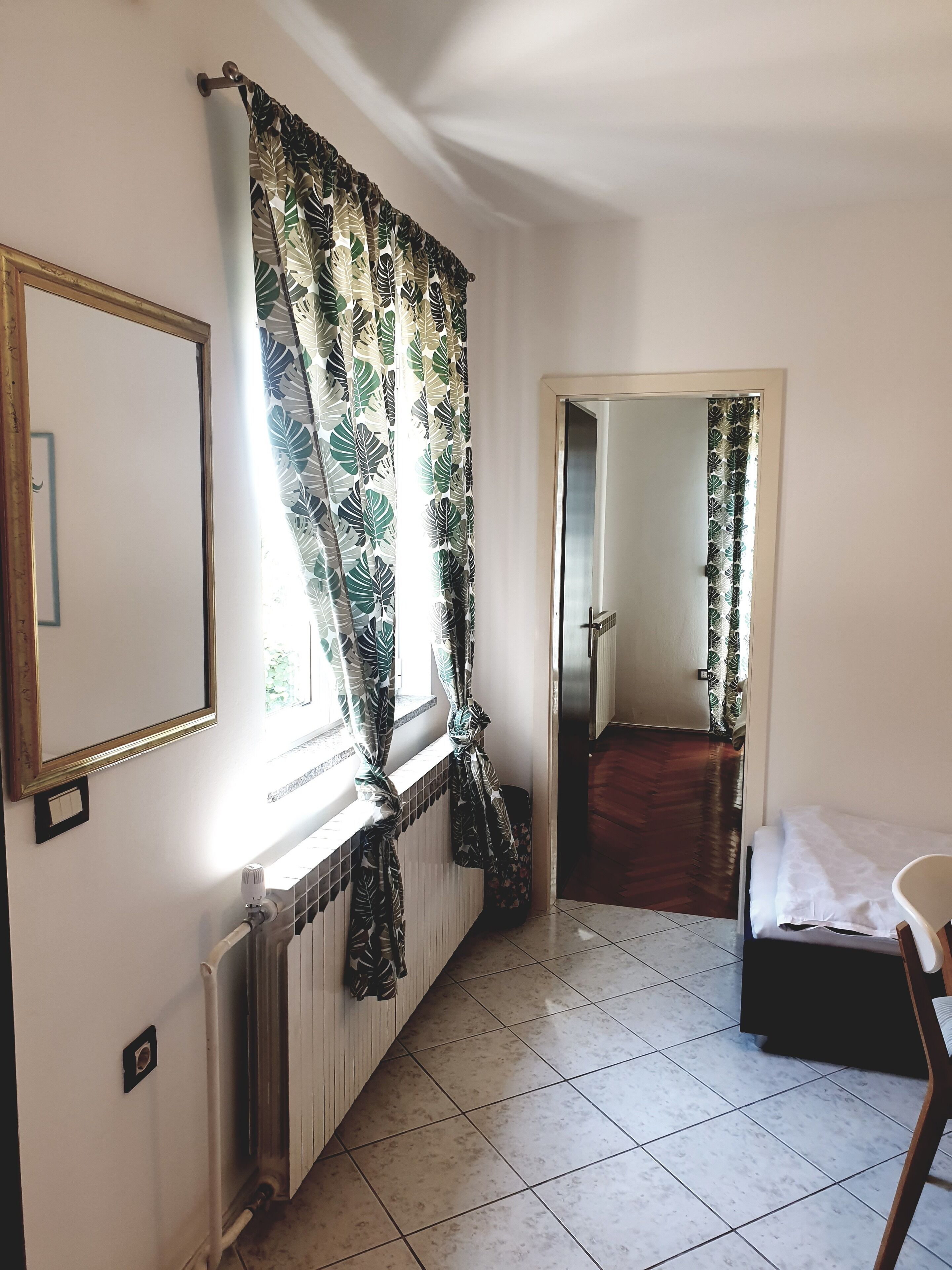 A room with a radiator, a window with curtains, a bed, and a door leading to another room.