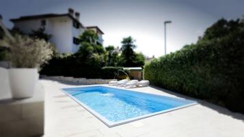 Una piscina con sillones de descanso, rodeada de un seto y un edificio al fondo.