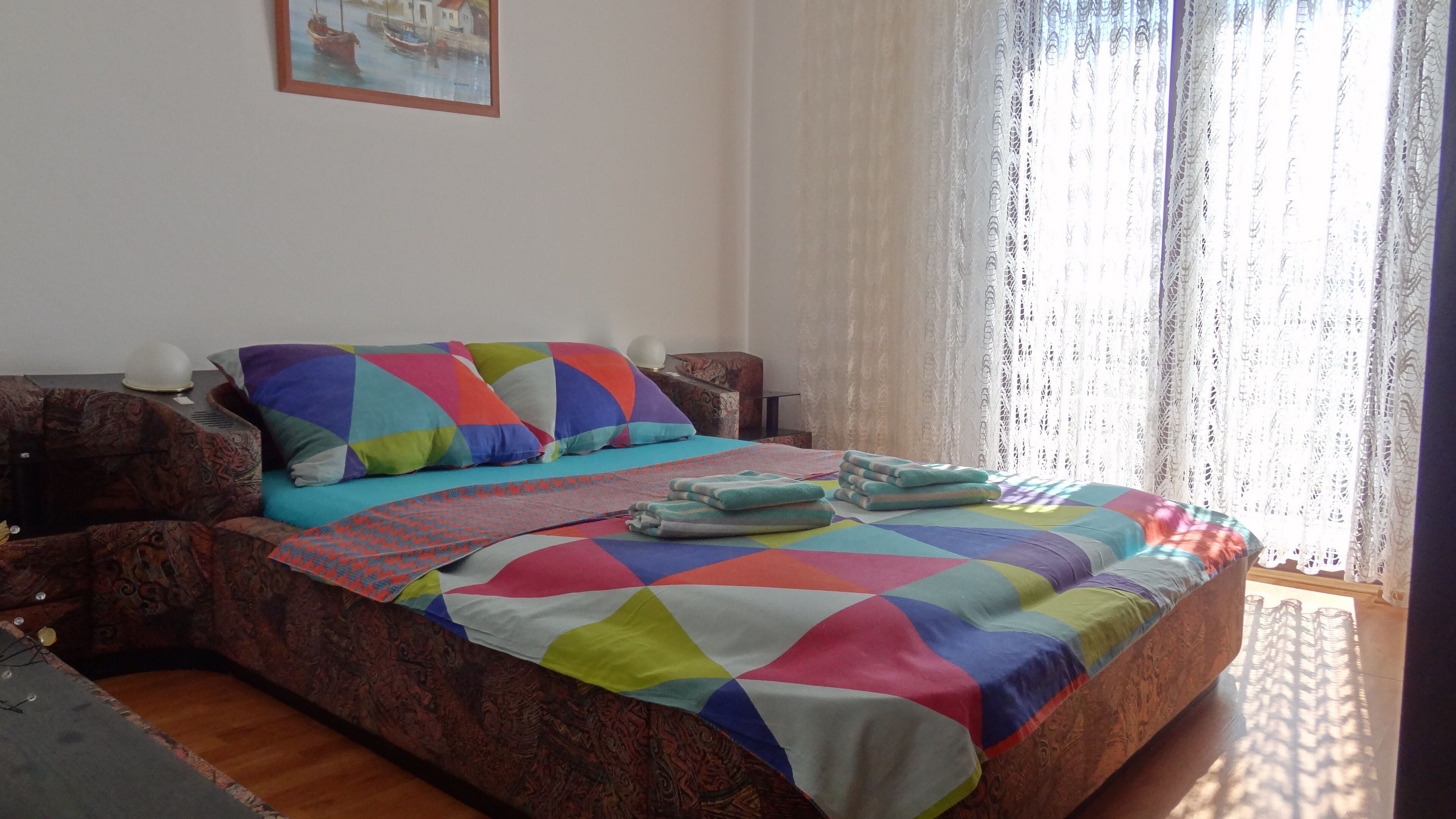 Una cama con un edredón colorido, un cabecero estampado y una ventana con cortinas traslúcidas.