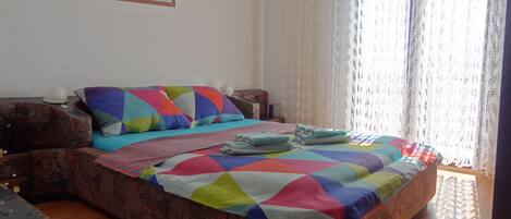 Una cama con un edredón colorido, un cabecero estampado y una ventana con cortinas traslúcidas.