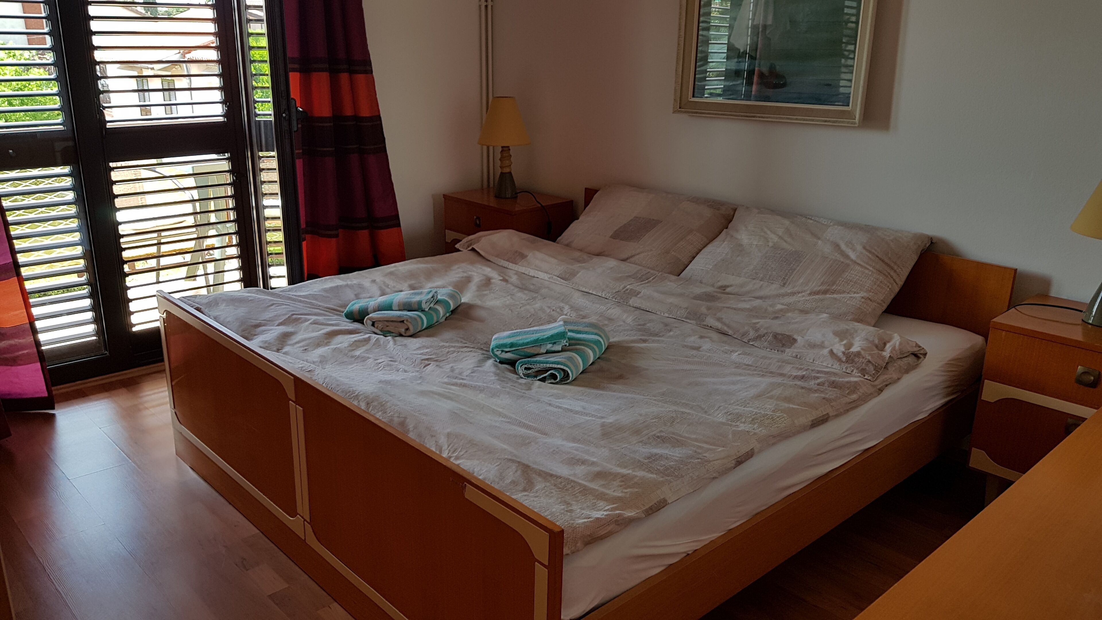 Una cama doble con estructura de madera, dos mesitas de noche, una lámpara y una ventana con persianas y cortinas.