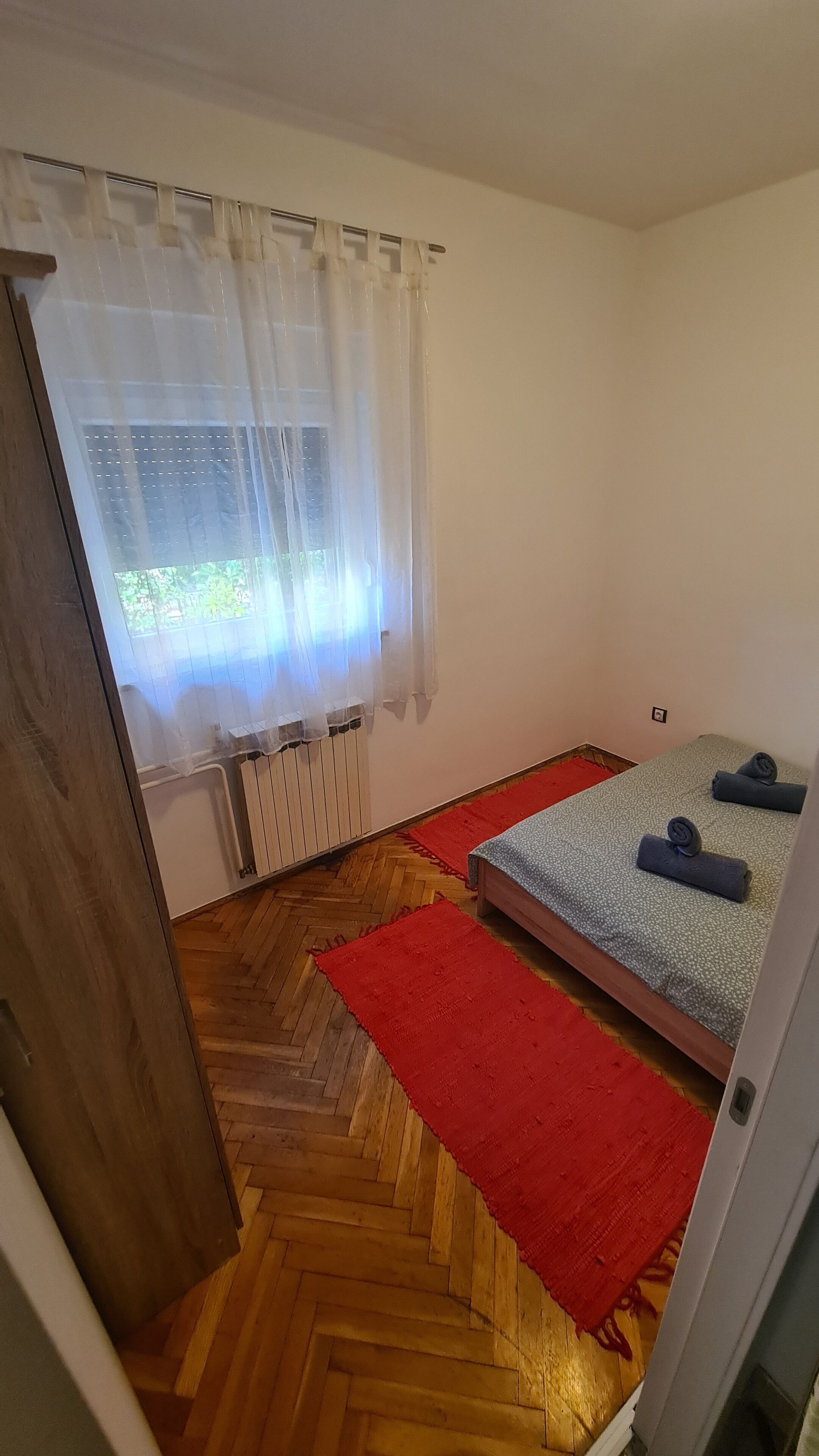 A small, tidy room with a wooden herringbone floor, a red rug, two beds with grey bedding, a window with sheer curtains, and a radiator.