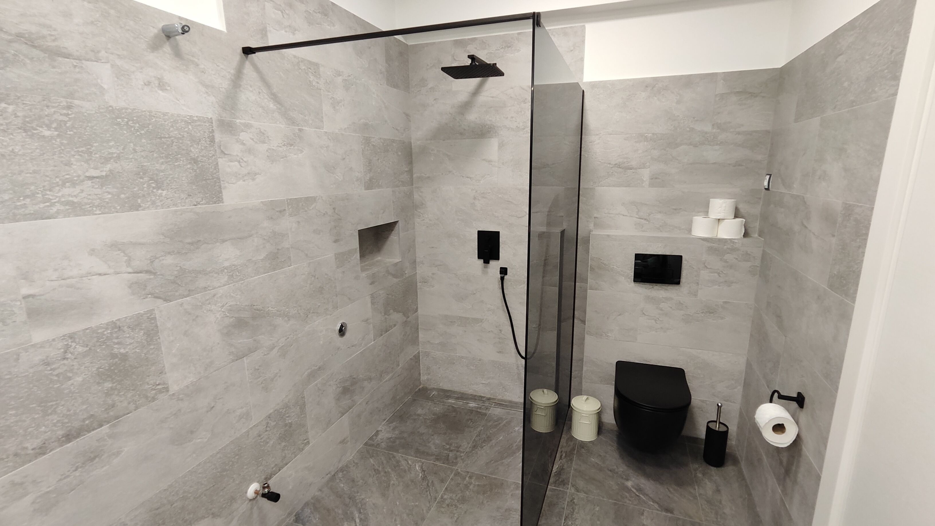 Een moderne douche ruimte met een glazen deur, grijze tegels, een handdouche, een toilet en een prullenbak.