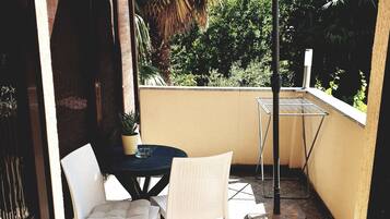 Une petite terrasse extérieure avec une table ronde, deux chaises en osier blanches et un grand parasol.