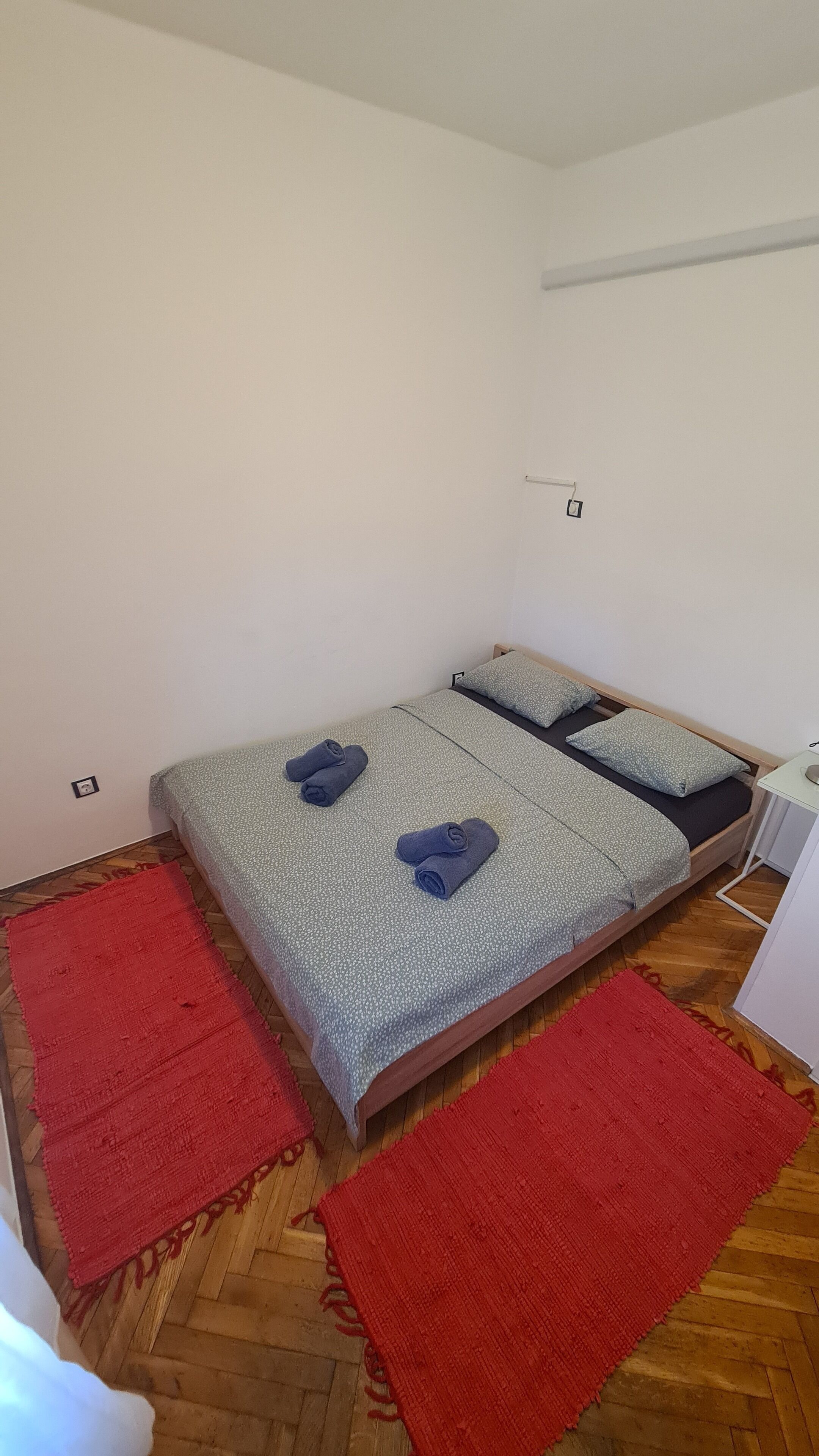 A small, neatly arranged bedroom with a bed, two pillows, and two rolled towels.