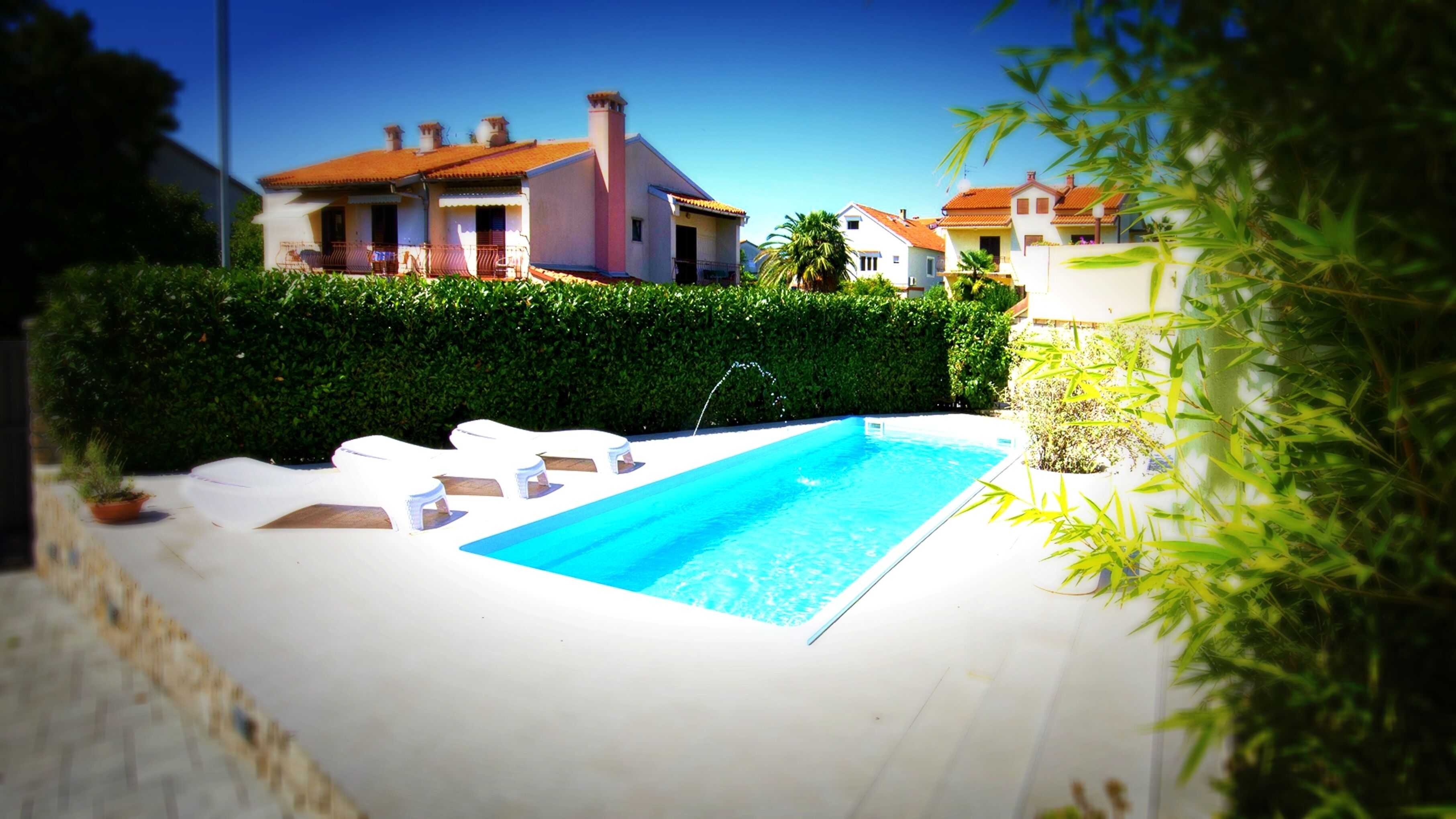 Una piscina con agua azul cristalina, rodeada de sillones blancos, un seto y una hilera de casas al fondo.
