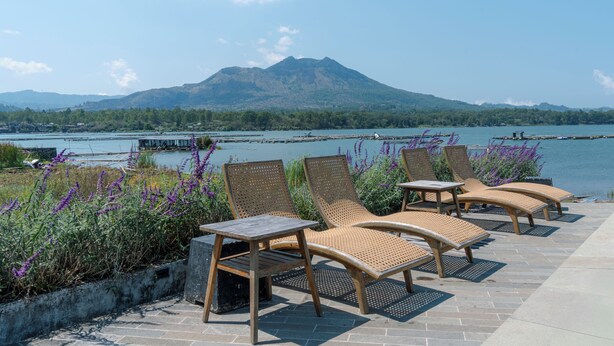 A terrace with lounge chairs and a table, overlooking a lake with mountains in the background. - Noya Hotel (Kintamani)