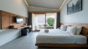 A modern hotel room with a large bed, a flat-screen TV, a wooden headboard, a small kitchenette, and a balcony with a view of the mountains. - Noya Hotel (Kintamani)