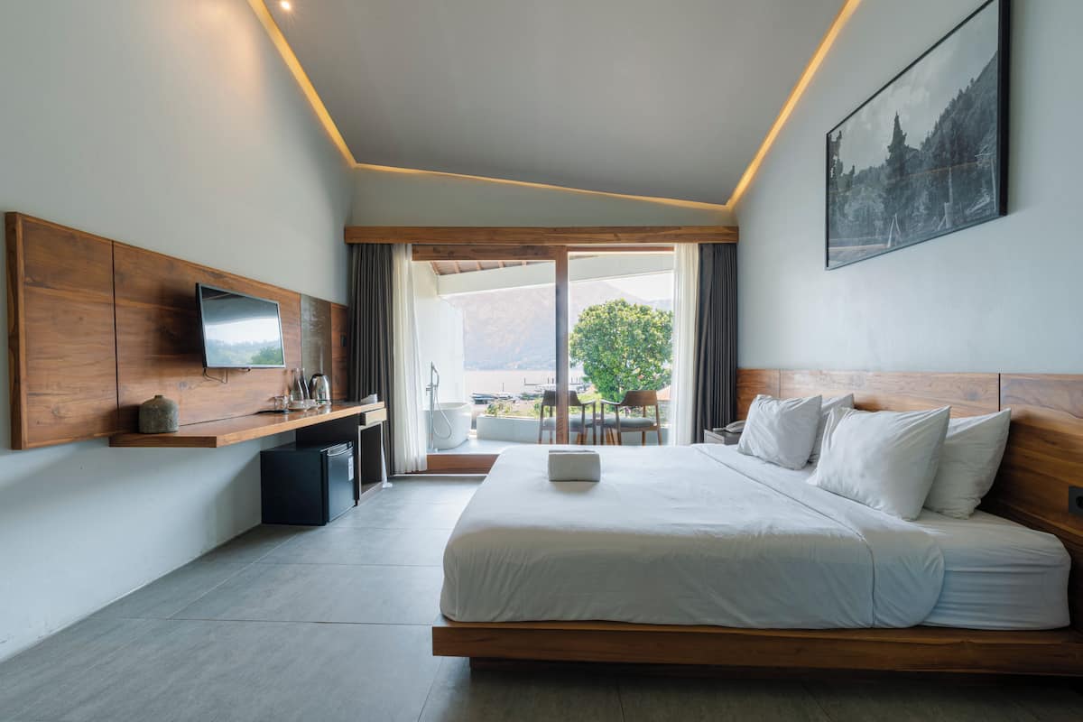 A modern hotel room with a large bed, a flat-screen TV, a wooden headboard, a small kitchenette, and a balcony with a view of the mountains.