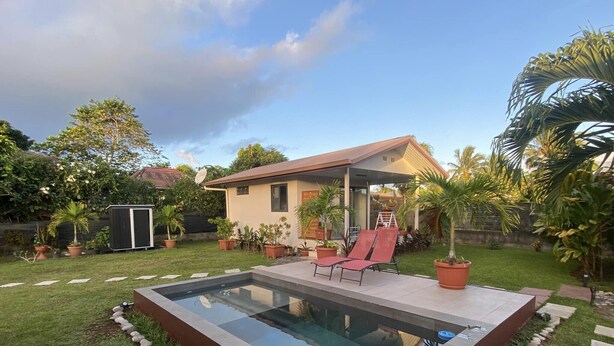 A modern house with a swimming pool, a patio with two red lounge chairs, and a small shed in the background.
