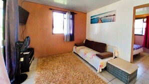 A room with a sofa, a small table, a window with curtains, a painting on the wall, and a television on a stand.