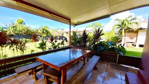 An outdoor patio with a wooden table and benches, surrounded by a garden with various plants and a view of the backyard.