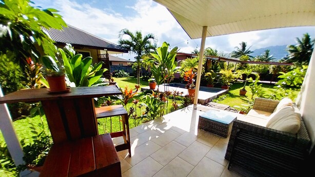 A covered patio with a table and chairs, surrounded by potted plants and a view of a swimming pool and mountains.