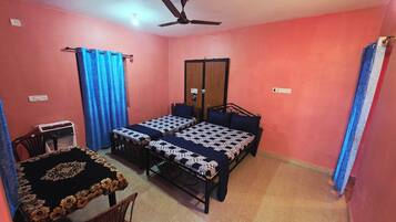 Una habitación pequeña con paredes rosadas, dos camas individuales, una mesa de madera, una silla, un ventilador de techo y una ventana con cortinas azules.