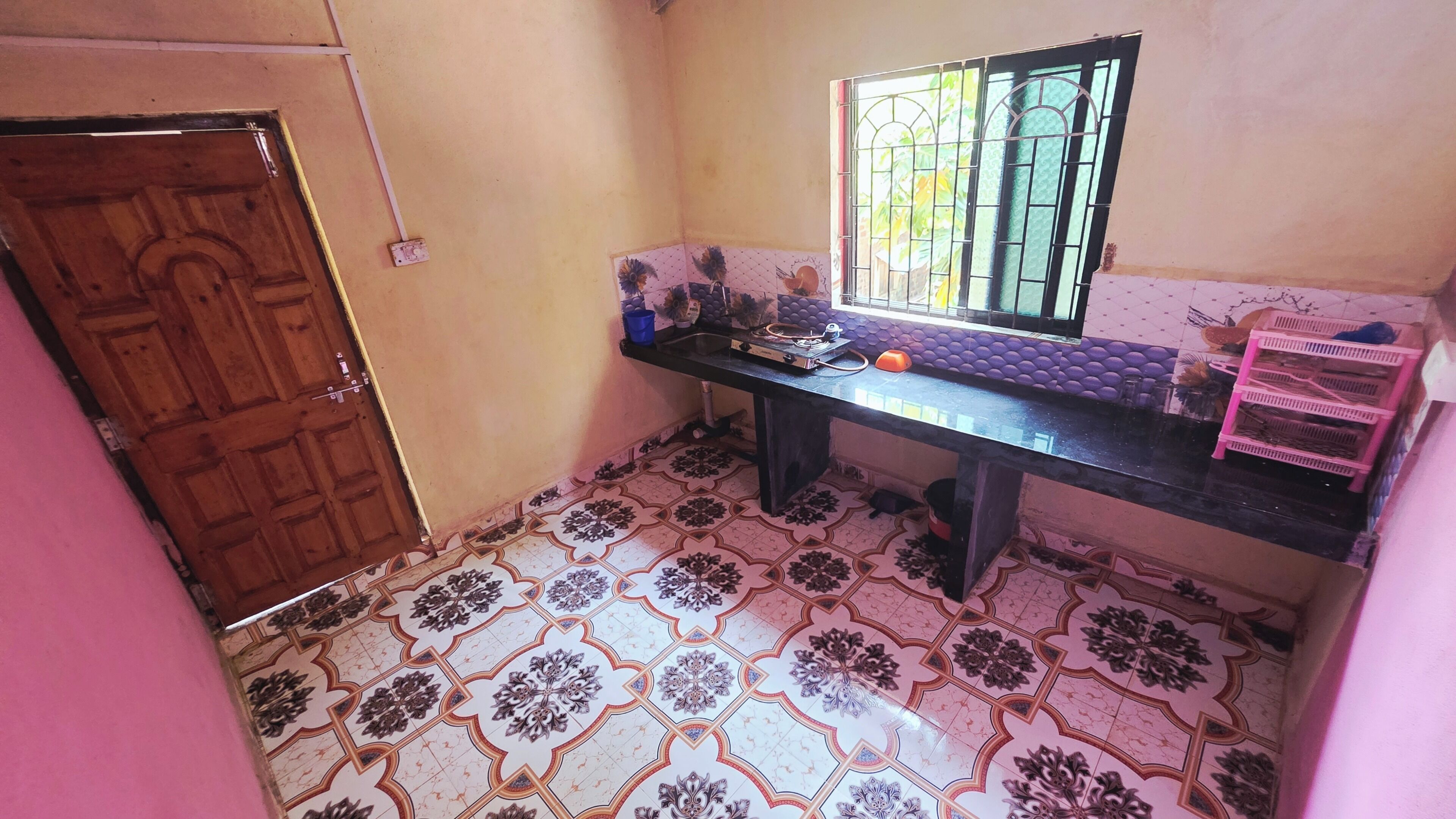 Uma cozinha com chão de azulejos, porta de madeira, janela com grades e prateleira rosa.