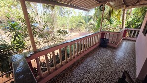 A covered balcony with a tiled floor, a metal railing, and a view of a garden with various plants and trees. - Vailankani guest house (Canacona)