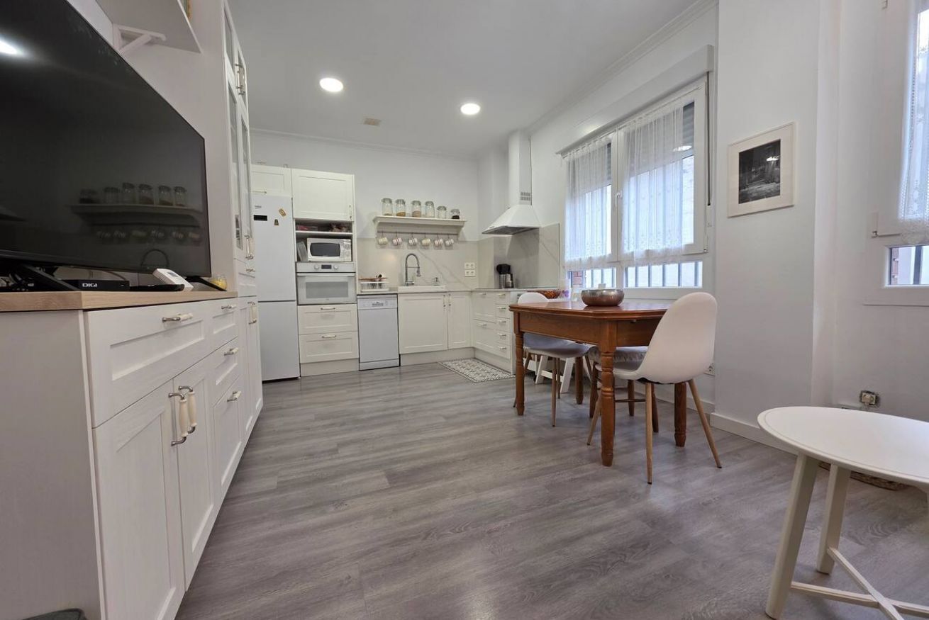 Een moderne keuken met witte kasten, een eethoek met een tafel en stoelen, en een groot raam met gordijnen.