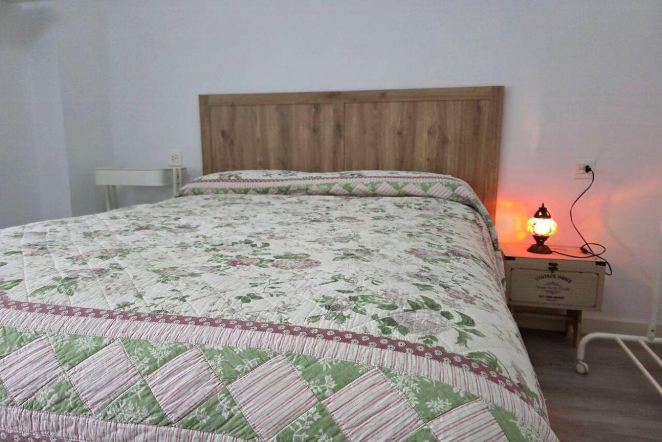 Una cama con colcha floral, cabecera de madera, mesita de noche con lámpara y toma de corriente.