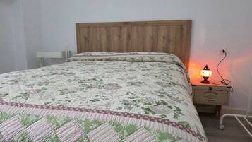 Una cama con colcha floral, cabecera de madera, mesita de noche con lámpara y toma de corriente.
