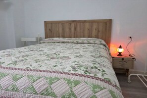 A bed with a floral quilt, a wooden headboard, a bedside table with a lamp, and a wall socket.