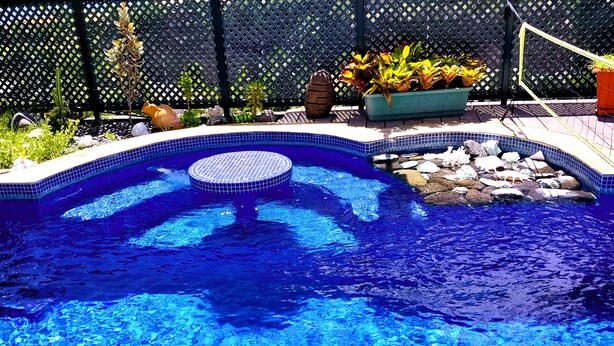 A swimming pool with a blue tiled edge, surrounded by a lattice fence, rocks, and potted plants. - TAHITI - Terevete Pool Studio (Arue)