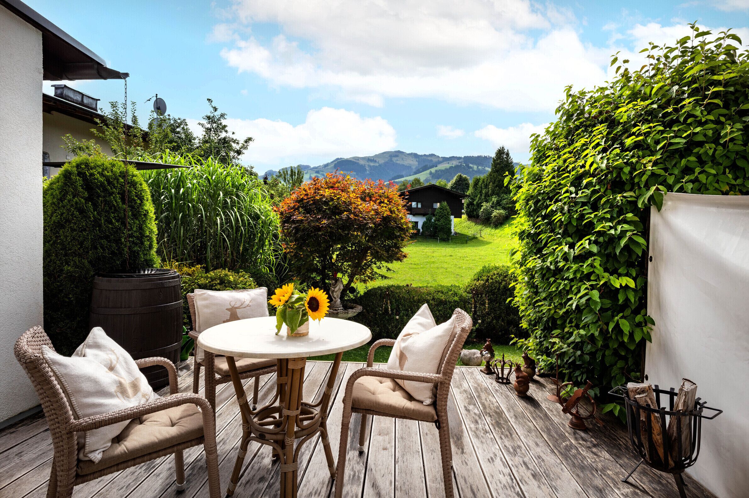 A wooden deck with wicker chairs and a round table, surrounded by greenery and a view of a house and hills.