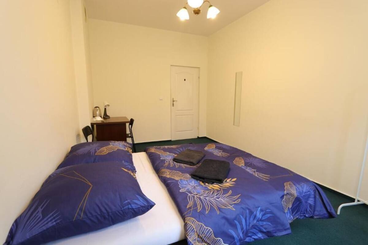 A small, single-bed room with a purple floral bedspread, a white mattress, and a small bedside table with a lamp.