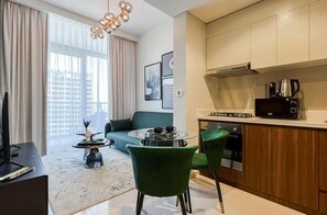 A modern living room with a green sofa, a coffee table, and a TV. There is a kitchen area with a stove, microwave, and cabinets. - Luxurious 1BR - Pool & Gym Access (Dubai)