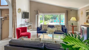 Un salon avec un canapé, un fauteuil rouge et un téléviseur. On y trouve une table en verre sur laquelle est posée une plante. La pièce est dotée d’une grande fenêtre avec des rideaux.