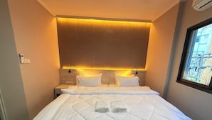 A neatly made bed with white linens and a quilted duvet, a bedside table with a lamp, and a window with a view of buildings. - Luck Hotel Huai Khwang MRT Ratchada (Bangkok)
