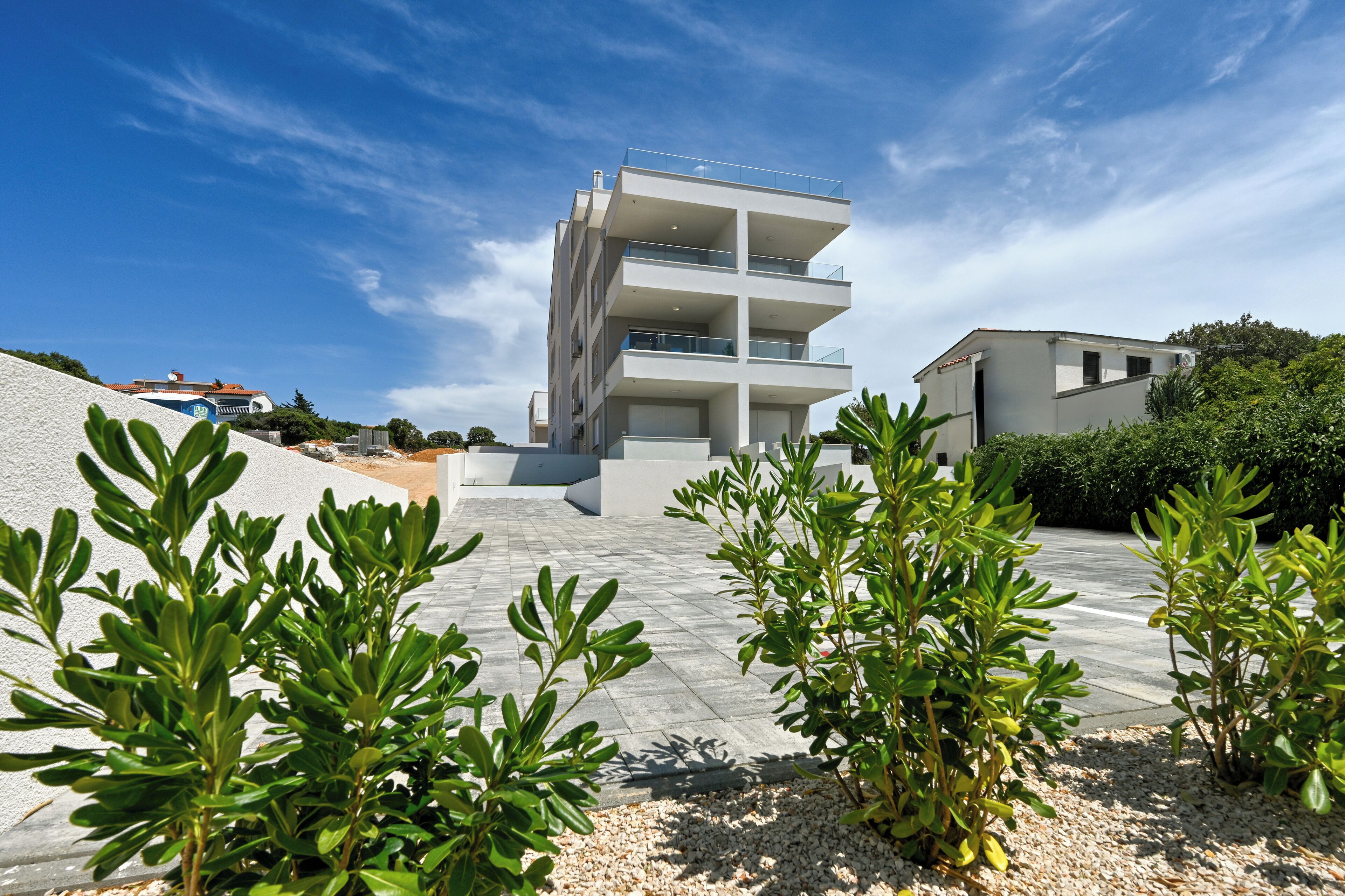 A modern multi-story building with balconies, surrounded by greenery and a clear sky.
