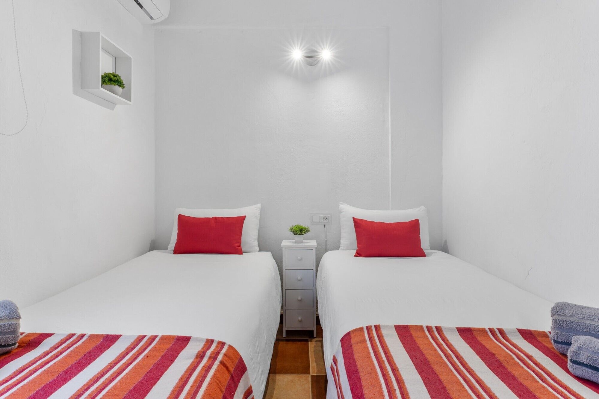 Una habitación pequeña y limpia con dos camas, una mesita de noche, un estante en la pared con una planta y una lámpara en el techo.