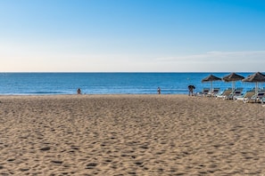 Ein Strand mit klarem Himmel, sandigem Ufer und einer Reihe aus Liegestühlen und Sonnenschirmen.