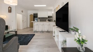 Una sala moderna con televisor de pantalla plana, un comedor con mesa de vidrio y una cocina con armarios blancos y un taburete de bar.