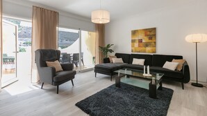 Una sala de estar moderna con una mesa de centro de vidrio, un sofá negro, un sillón gris y un amplio ventanal con cortinas.