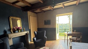 Une pièce avec un plancher en bois, un grand miroir, deux chaises, une petite table et une vue sur la verdure à l’extérieur.