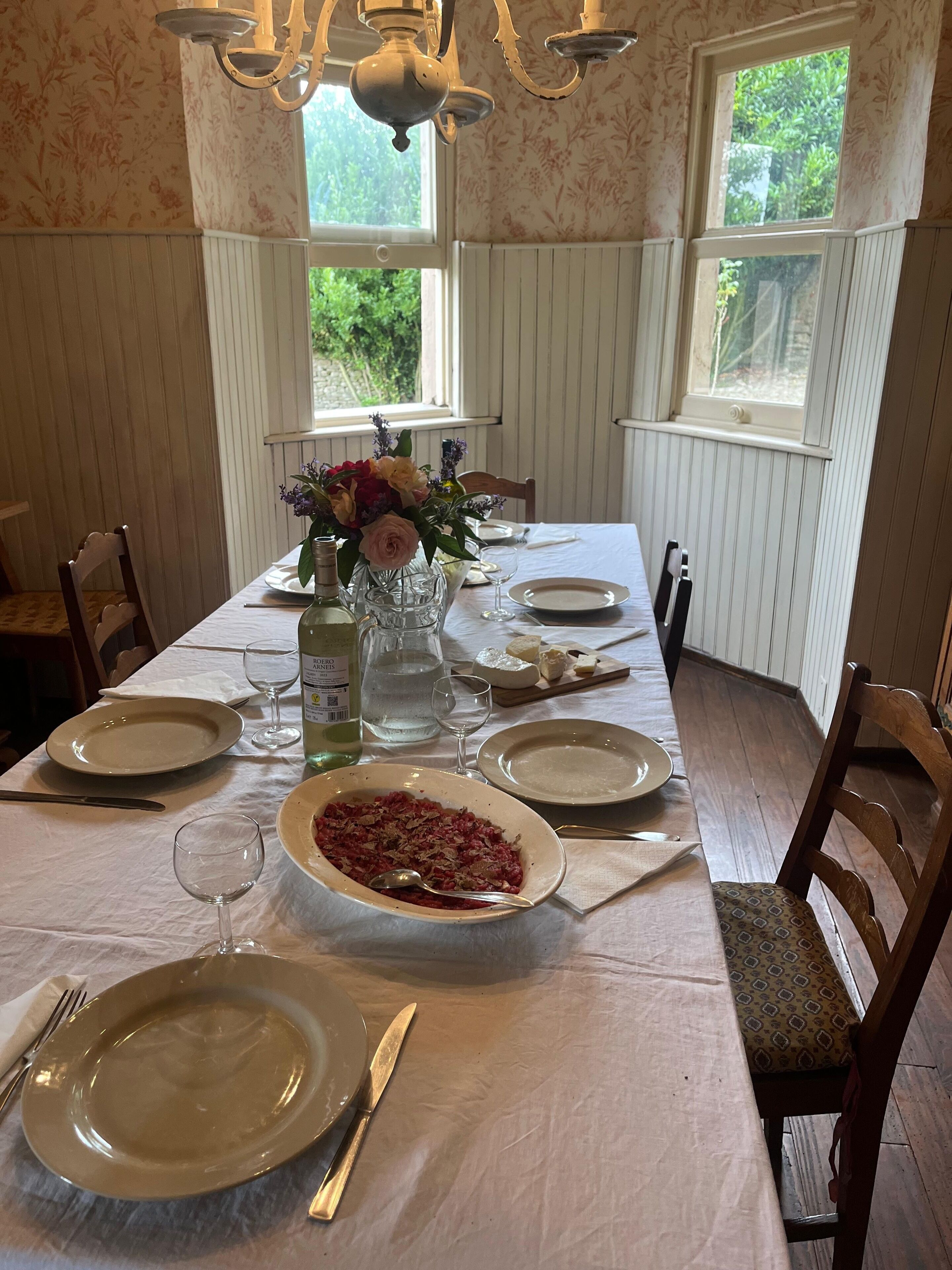 A dining table set for four with a central dish of food, wine glasses, and a vase of flowers.