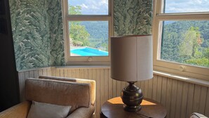 A room with two windows, a sofa, a table with a lamp, and a view of a pool and forest. - Villa with garden and pool. Unmissable view of the Langhe 200m from the village. (Bossolasco)