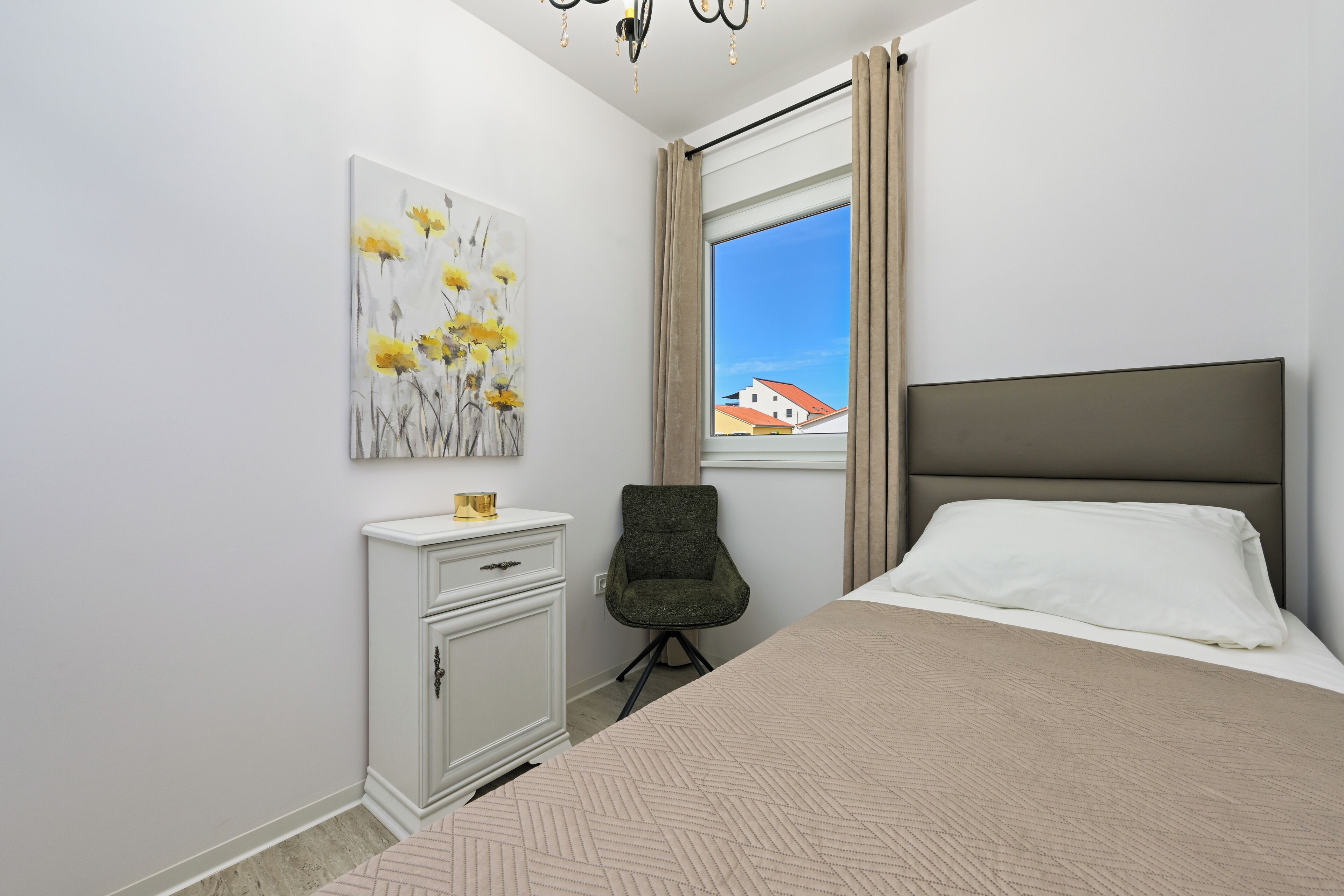 Un dormitorio con cama, mesita de noche, silla, ventana con cortinas y un cuadro colgado en la pared.